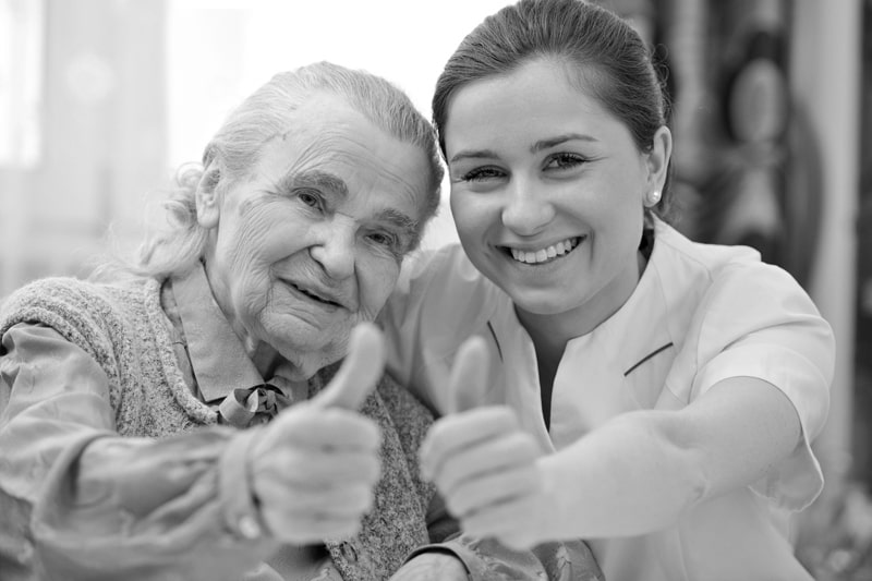A warmhearted nurse and an elderly woman with alzheimer's smiling while each giving a thumbs up